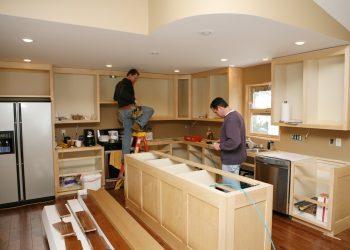 Cabinet maker installing custom made kitchen island and cabinets, while electrician installs in-cabinet lighting. 
Very shallow DOF quickly drops off to nice soft focus.